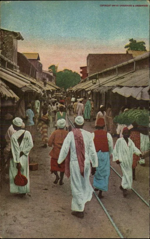 Hand-coloured vintage photo of a busy market street with people in kanzus